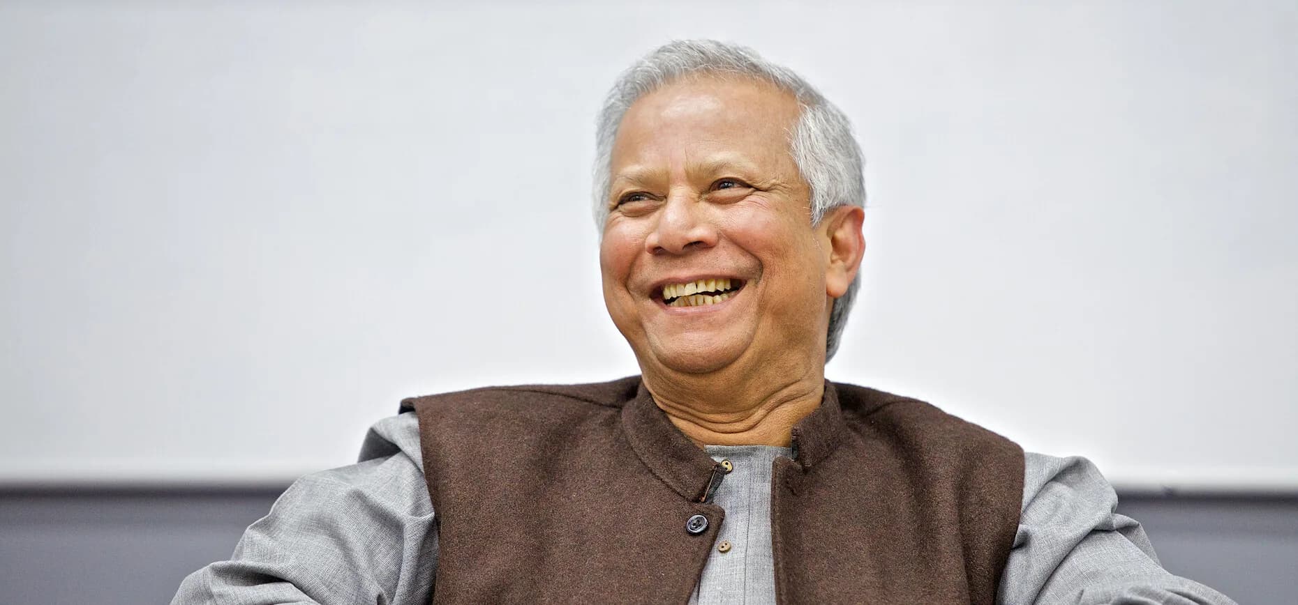 Professor Muhammad Yunus smiling, seated in a grey kurta and a brown waistcoat